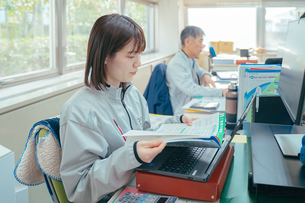 デスクで書類とPCに向かう女性社員。奥に男性社員。