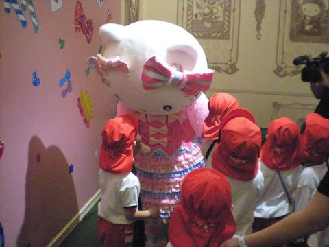 Hello Kitty with children in red hats, smiling and 年少組　散歩 enjoying time together inside.