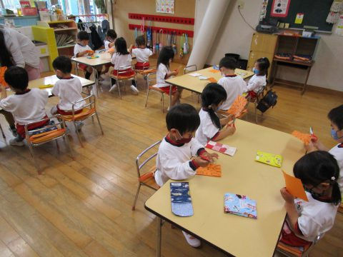 幼稚園で工作をする子供達。オレンジ色の用紙で作品作りに集中。始業式、楽しそう。