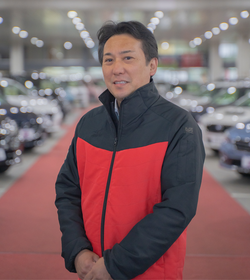 車を背景に立つ、赤と黒のジャケットを着た笑顔の男性社員
