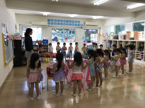 先生と水着姿の子供達が並んでいる様子。７月のお誕生日会。保育園の教室で水遊びの準備。