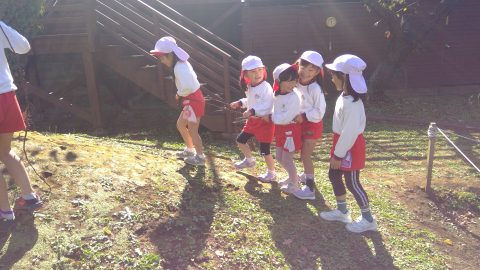 幼稚園児が階段を登る様子、白い帽子と赤いスカート姿です。３月【年中】体育指導、楽しそう。