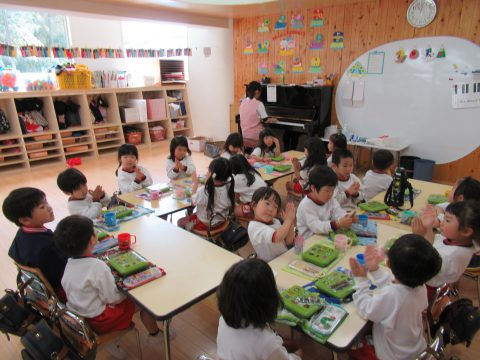 保育園の子供達が食事中。教室でピアノ演奏、黒板、棚。日本の幼稚園の風景。