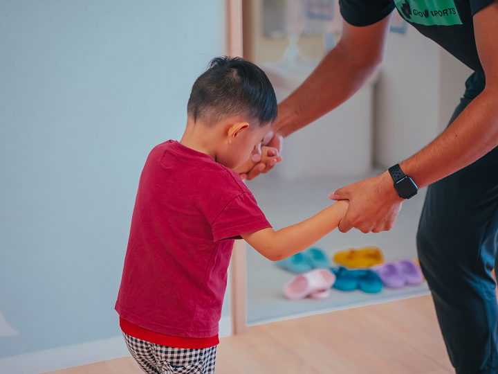 男の子が手を引かれ歩く、保育士が手伝う、元気よく歩く、背景に靴