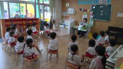 保育園の教室で先生が絵本を読む。1月【年少々】節分製作、子どもたちが座っています。