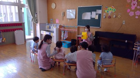 保育士と園児たち、教室にて。先生は黄色い紙を持つ。3月【年長】ピクニックランチ・外遊び。