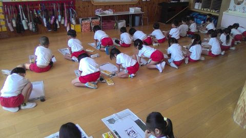 幼稚園児が床で絵を描いています。12月【年中】今週の様子、集中して制作活動中。木製の床の教室です。