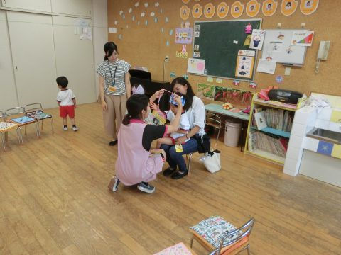 保育園の教室で、子供たちが楽しむ様子。先生と保護者も一緒。7月のお誕生日会、楽しい雰囲気。