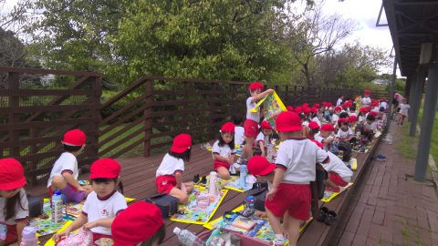 園児たちが野外活動中に座って、絵本を見ている様子です。5月【年少】今週の様子 青空の下。