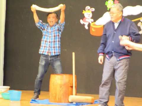 男性が餅を持ち上げ、餅つきをする様子です。年長組 参観日、背景に装飾あり。