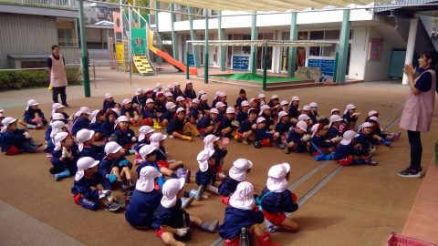 保育園の子供達と先生達。園庭に座って話を聞いています。5月【年少】今週の様子です。