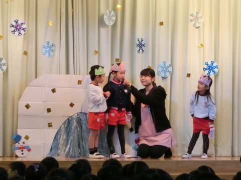 ステージ上の子供達と先生。子供達は冠を被る。始業式、背景には雪の結晶。