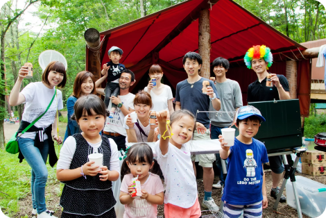 赤いテントの前で飲み物を持ち笑顔で並ぶ子供と大人のグループ