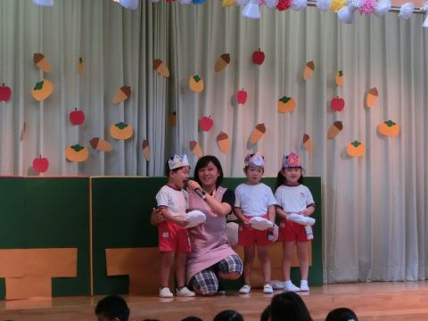 先生と園児たち、舞台で笑顔。秋の飾り付け。 英語 ハロウィンをテーマにしたイベントです。