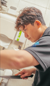 男性が真剣な表情で作業中、手に包丁を持つ。 背景はオフィス。株式会社ao