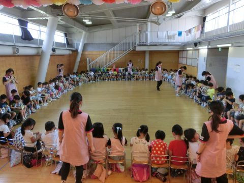 園児たちが輪になって座り、先生たちが見守る様子。年長組　参観日、保育園での活動。