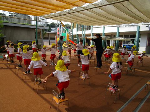 園児たちが椅子で運動、先生が見守る様子。5月【年中】英語指導、屋外の遊具も見える。