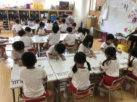 教室で音楽の授業、子供たちが楽譜を書く様子。全員白いシャツと赤いズボン。