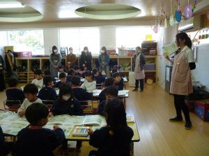 園児たちと先生たち、教室で何かを読む。始業式、子供たちが真剣に聞いている様子。