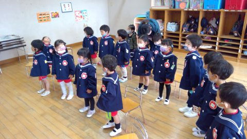 園児たちが整列し先生の話を聞いている様子。12月【年少】大掃除、背景は教室、冬の制服。