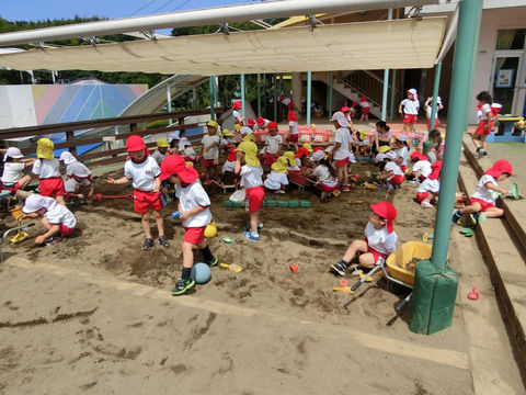 砂場で遊ぶ園児たち、赤い帽子と体操服姿。運動会中の砂遊びの様子です。楽しそう