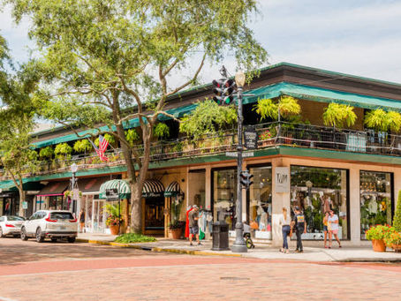 People walk by shops with green awnings on a leafy street corner. Hanging plants are visible on the balcony, along with a U.S. flag.