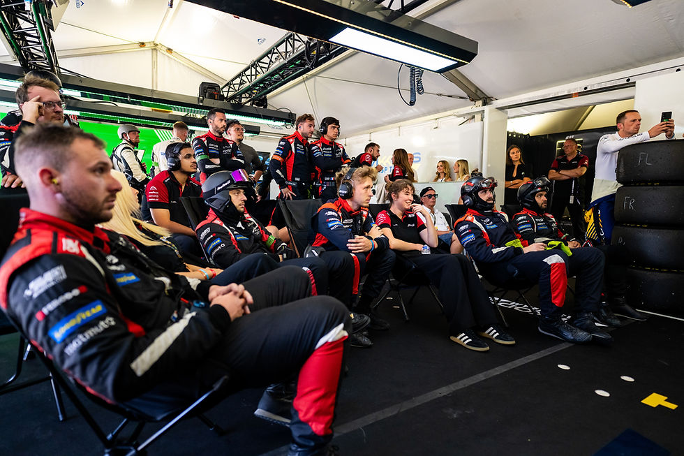 ASP team members in black-red uniforms watching the race in a pit garage; helmets on, with FR and FL marked on stacked tyres | Credit: DPPI
