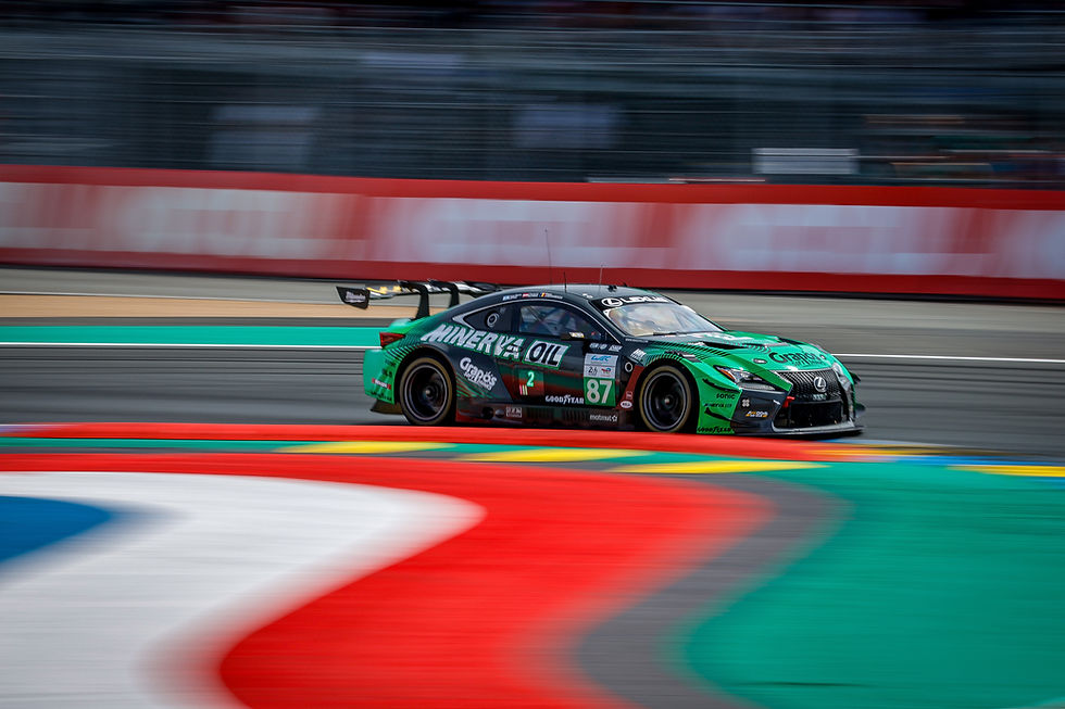 Green-and-black Lexus race car No.87 speeds past a blurred track, with red, white, and teal curbs and sponsor logos visible. | Credit: DPPI / Paulo Maria