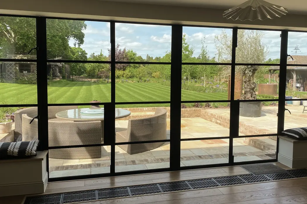 Full-height steel-framed glazing looking out from a living space onto a paved terrace and striped lawn.