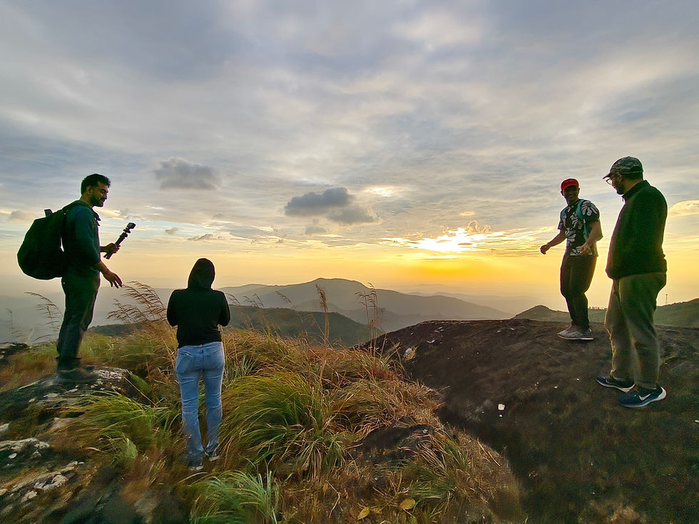 Four people stand on a grassy hilltop at sunset, with one holding a camera. The sky is cloudy, and distant mountains are visible.