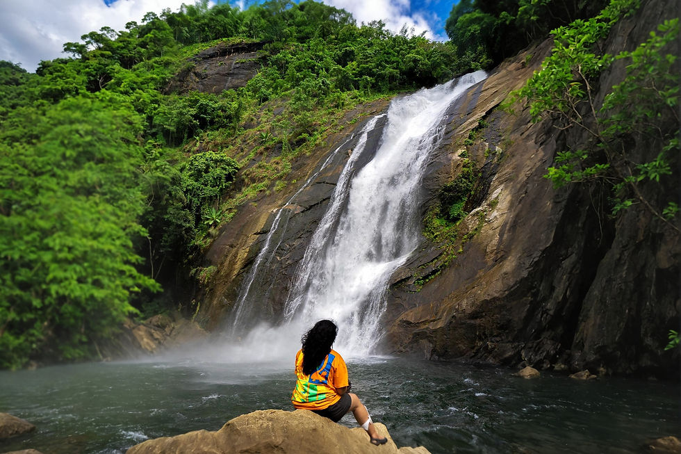 Person in orange shirt sits on a rock facing a tall waterfall amidst lush greenery under a blue sky, creating a peaceful scene.