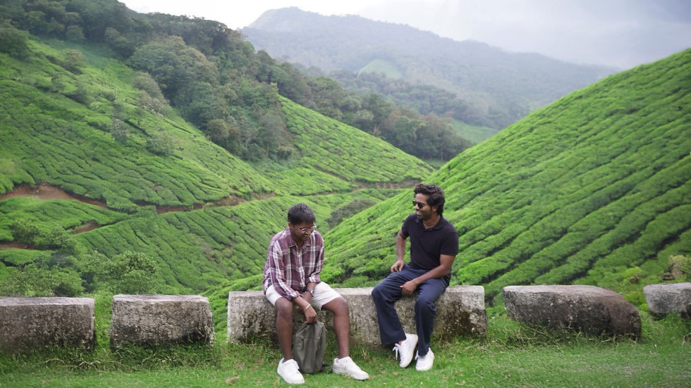 Two men sit on stone barriers, laughing in a lush green tea plantation with rolling hills. The sky is overcast, creating a serene mood.