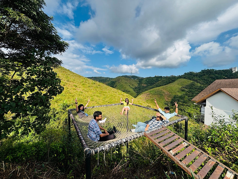 Five people relax on a net over lush hills, with arms raised in joy. A small building and a blue sky with clouds are in the background.