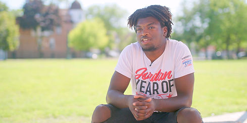 A young man sits outdoors on a bright day, wearing a white t-shirt with red and black text, speaking while looking towards the camera. The background is blurred, featuring a grassy field and trees, providing a calm and natural setting