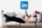 A relaxed man in casual attire reclines on a sofa, wearing headphones and holding a tablet stylus, with his arm extended towards a floating LinkedIn logo above him, symbolizing professional networking and content engagement on the platform