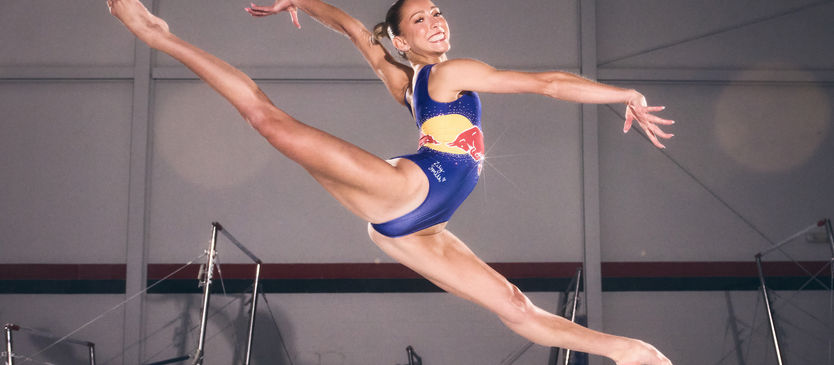 Athlete Lily Smith in the middle of a split leap in her red bull leotard smiling. 