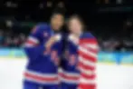 Athletes Laila Edwards and Hilary Knight of Team USA at the Olympics on the ice, holding their gold medals with an american flag wrapped around them.