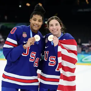 Athletes Laila Edwards and Hilary Knight of Team USA at the Olympics on the ice, holding their gold medals with an american flag wrapped around them.