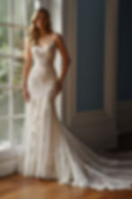 A bride in an elegant, lacy white wedding dress stands by a large window. Sunlight highlights intricate details, creating a serene mood. MARTIN THORNBURG, MON CHERI BRIDALS, BOCHIC BRIDAL BOUTIQUE, CINCINNATI OHIO, READING BRIDAL DISTRICT
