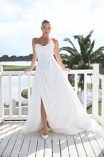 Woman in a white strapless wedding gown stands on a sunlit deck by water and palm trees, gazing at the camera.