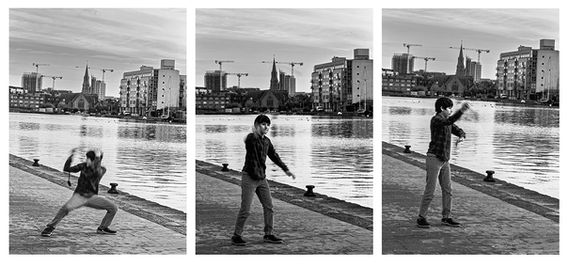 Trittico fotografico in bianco e nero che ritrae un giovane ai Grand Canal Dock di Dublino nel novembre 2025. La sequenza cattura tre momenti di un lancio con la fionda: la preparazione carica di tensione, la mira stabile e il rilascio finale. Sullo sfondo, le architetture moderne e le gru dei Docks creano un contrasto grafico tra l'azione umana e il paesaggio urbano.