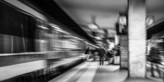 Fotografia artistica in bianco e nero di un treno in movimento sfuocato in una stazione, con un uomo fermo sulla banchina in forte contrasto visivo.
