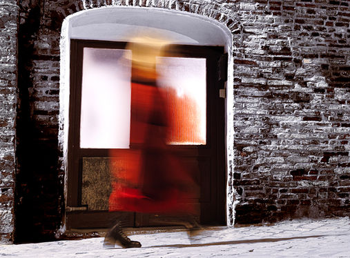 Fotografia d'arte in tempi di esposizione lunghi con una figura femminile sfocata in rosso che attraversa una porta, accanto a un muro in mattoni. Massimo Cicconi Fotografia.