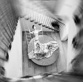 Fine art black and white photograph published on 1x. A zenithal perspective of a monumental staircase with a dynamic radial effect framing a surreal figure on a ladder.