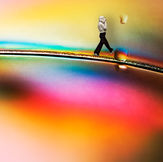 Fotografia d'arte creata con polarizzazione incrociata e manipolazione digitale. Una piccola silhouette umana cammina in equilibrio su una linea sottile e scura che attraversa un paesaggio astratto di colori iridescenti, arcobaleni e texture fluide, simboleggiando l'interazione umana con le leggi della fisica