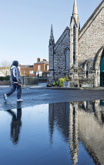 Visione specchiata della cattedrale di Rathmines dopo la pioggia. Il riflesso sull'asfalto bagnato duplica le linee gotiche della chiesa e la sagoma di un pedone