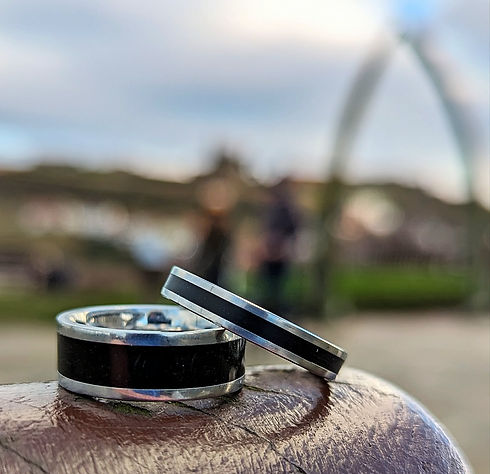 Two wedding memorial ashes rings containing real Whitby Jet, resting on a stone ledge in front of the iconic Whitby whale bones arch, with a soft, blurred Whitby town background.