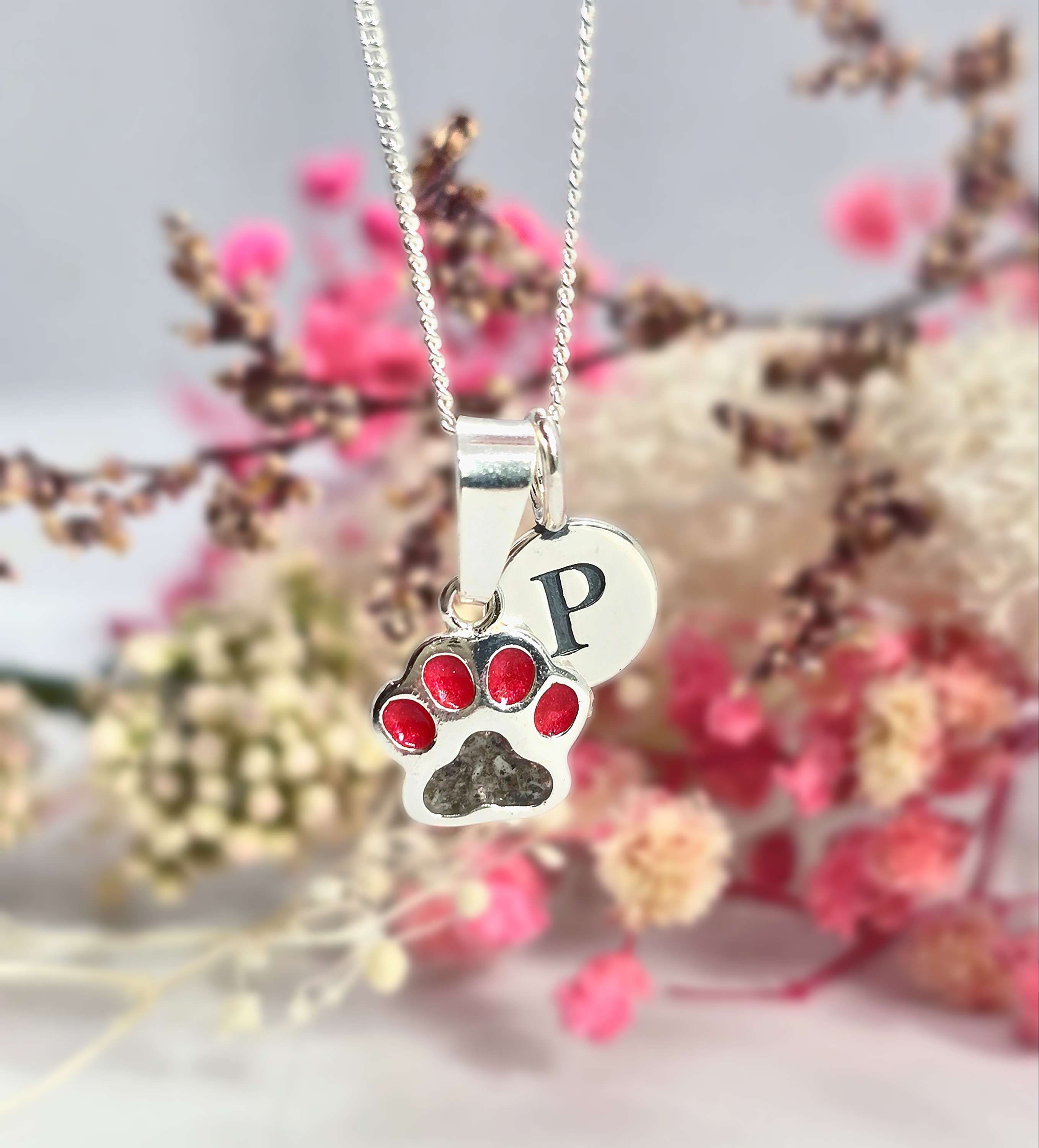 Sterling silver pawprint ashes necklace with colour-filled toe beans and initial charm, displayed on floral background.