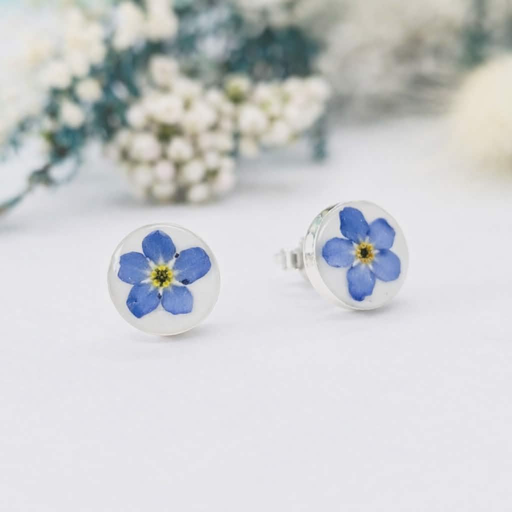 Forget-me-not breastmilk earrings, 8mm studs with pressed blue flowers over white breastmilk resin, shown with dried floral b