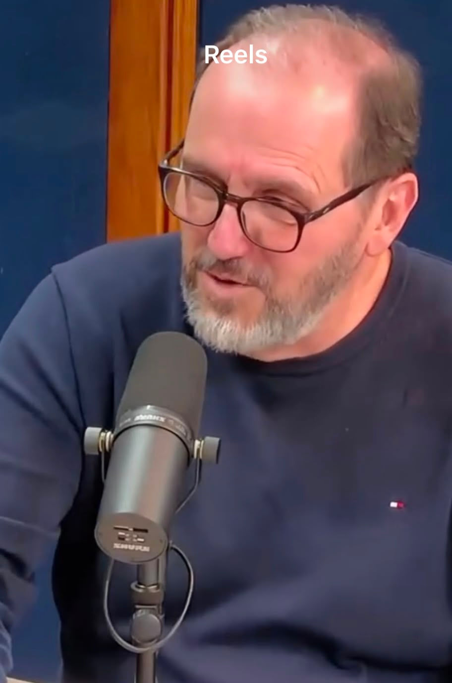 Homem barbudo de óculos fala ao microfone em estúdio azul; texto Reels no topo.